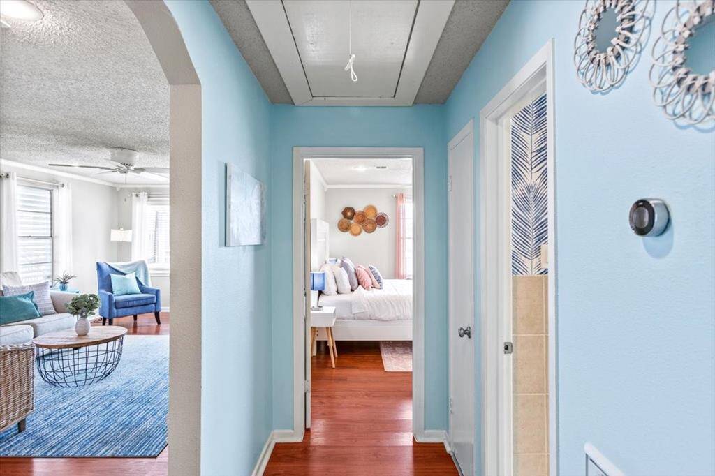 This is a bright and airy hallway featuring light blue walls and hardwood flooring, leading to a cozy bedroom. The living area is visible on the left, showing a comfortable and stylish space with modern furnishings and ample natural light.