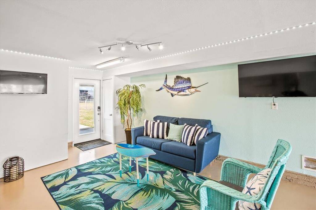 This is a bright living space with a tropical theme, featuring a comfortable seating area with a blue sofa and two wicker accent chairs, LED ceiling lighting, a mounted flat-screen TV, and a decorative marlin on the wall. The room has a fresh feel with a light blue accent wall and opens directly to the outdoors through a glass door, inviting natural light.