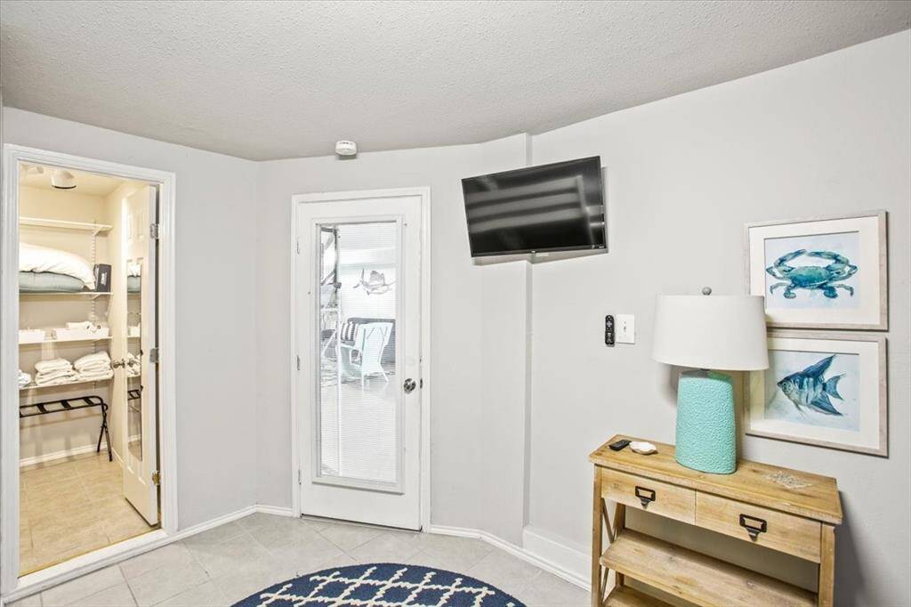 Door to outside living space. This is a cozy corner space featuring a glass door that leads to an outdoor area, a small wooden console table with a lamp, and two marine-themed pictures on the wall. A flat-screen TV is mounted opposite the door, and the room opens into a closet or storage area with shelving. The decor suggests a beach or coastal theme.
