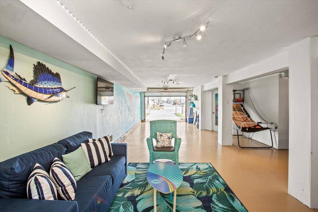This is a spacious, well-lit living area with a tropical theme, featuring eclectic decor, track lighting, and a flowing layout that leads to additional rooms. The space includes a comfortable seating area and artistic touches, such as a wall-mounted fish sculpture.