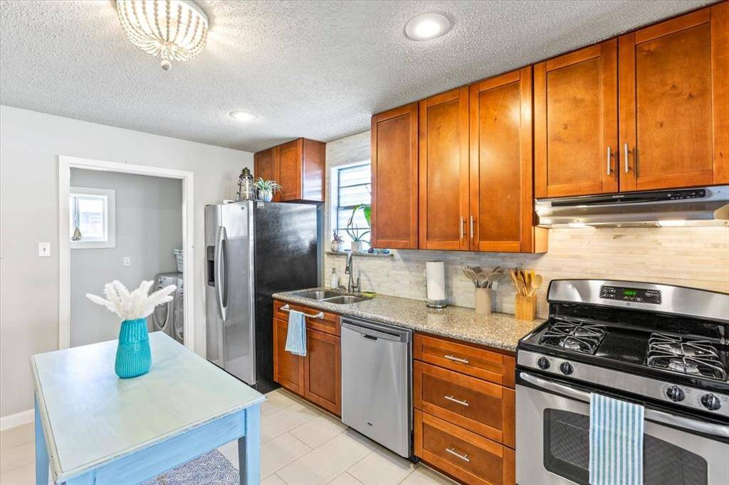 This kitchen features modern stainless steel appliances, ample wooden cabinetry, a gas stove, and a central island providing additional workspace. The room is well-lit, with a stylish light fixture and natural light coming through a window. It's finished with tile flooring and a neutral color scheme.