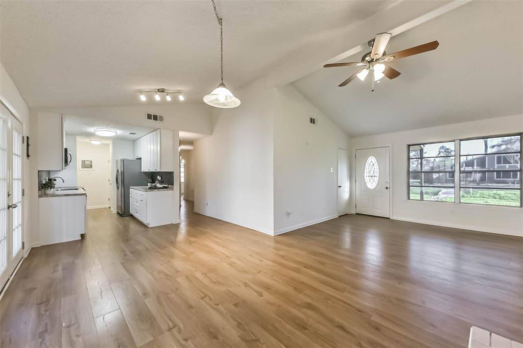 This photo showcases an open-concept living area with a high ceiling and ceiling fan, featuring engineered wood flooring. The space includes a kitchen with modern appliances and white cabinetry, adjacent to a dining area with glass French doors leading outside. Natural light floods the room through large front windows.