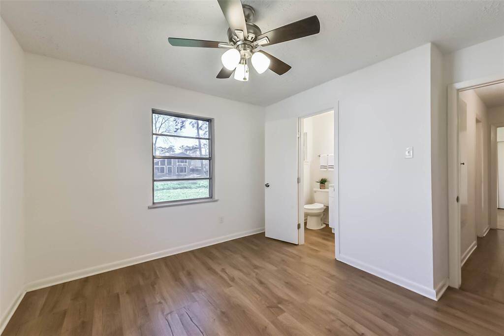 Bright bedroom with wood flooring, a ceiling fan, and an en-suite bathroom. The room features a window providing natural light and a view of the outdoors.