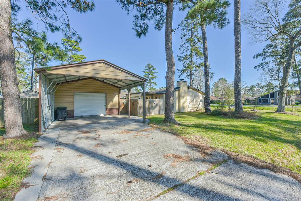This property features a spacious driveway leading to a covered carport and garage. It's surrounded by tall trees, providing a serene, wooded setting. The home is situated on a grassy lot in a quiet neighborhood, offering privacy and a natural landscape.