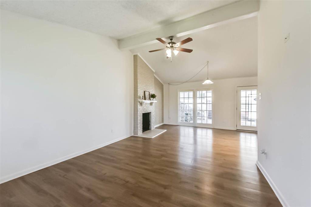 This spacious living room features vaulted ceilings, a ceiling fan, and a cozy brick fireplace. The room is brightened by large windows and a glass door leading to the outside, with warm wood flooring enhancing the inviting atmosphere.