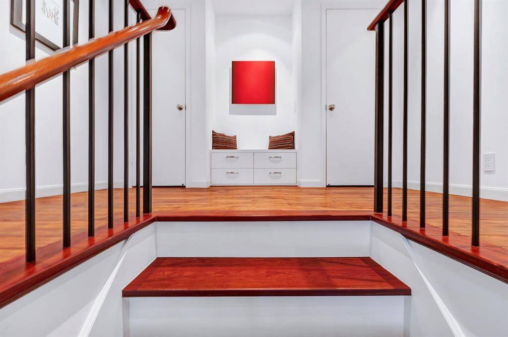 Exemplifying a high level of craftsmanship, the staircase ascends with precision, featuring solid Brazilian Cherry wood treads, white risers, and flat black wrought iron balusters for a striking contrast. A matching cherry handrail seamlessly guides the ascent to the upstairs landing, where a symmetrically framed built-in seating nook is flanked by dual closets, blending functionality with architectural balance. Thoughtful detailing and precise alignment enhance this refined design.