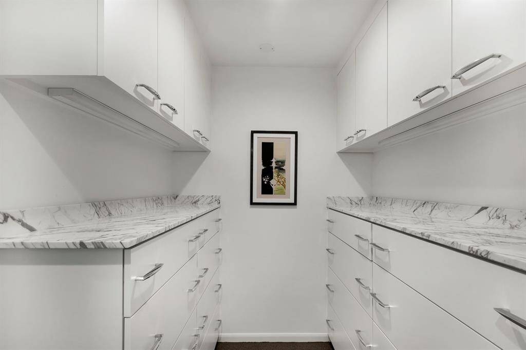 Adjacent to the owner’s bath, this spacious walk-in closet features built-in dressers with luxurious Arabescato white marble countertops. As part of the 2007 owner's bath renovation, a closet addition (not shown) expanded the space with a left entryway leading to an expansive walk-in closet with built-in shelving, ample hanging rods, and integrated storage for a seamless, organized design.