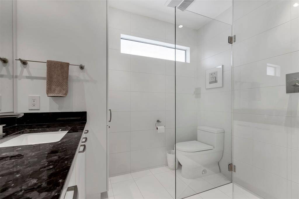 A frameless glass walk-in shower enhances the open feel, while the fluted glass transom window allows natural light to filter through, maintaining privacy. Thoughtful details, such as the understated tilework and fixtures, contribute to the bathroom’s sophisticated yet practical aesthetic.