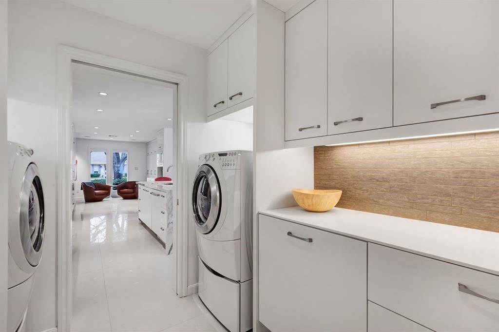 This luxury laundry suite combines a refined design with high functionality, featuring an interlocking porcelain tile backsplash, Corian solid surface countertops, and custom cabinetry with solid surface door and drawer fronts, mirroring the kitchen’s style. Undercabinet lighting enhances the polished, modern look. Conveniently situated between the kitchen and attached garage, this thoughtfully designed space offers both efficiency and elegance for everyday living.