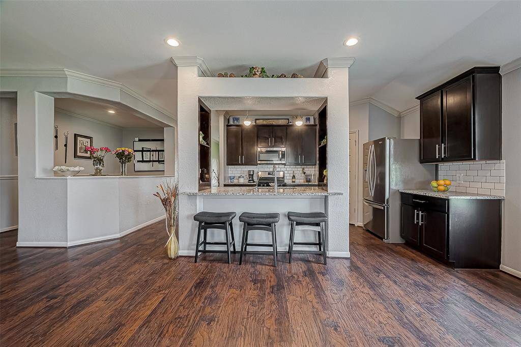 This photo showcases a modern kitchen with dark wood cabinets, stainless steel appliances, and a breakfast bar with seating space for three. The breakfast bar features elegant laminate wood-like flooring, recessed lighting, and a stylish tile backsplash. Notice the pretty built-in shelves.
