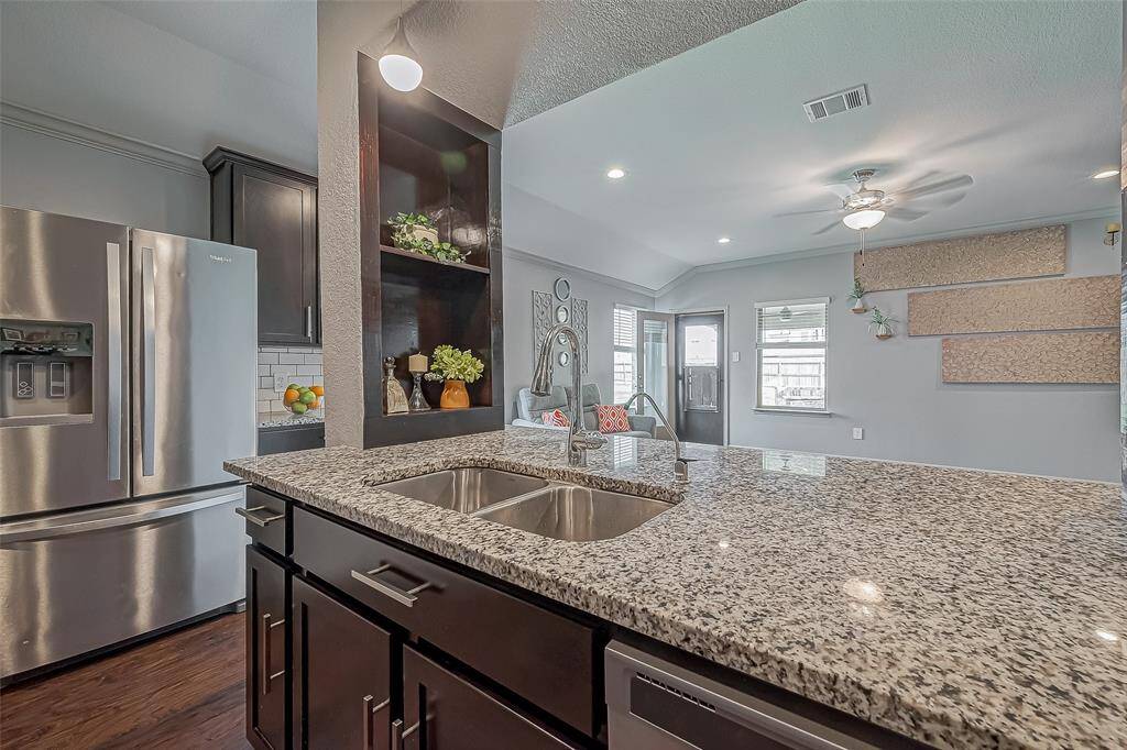 This photo showcases a modern kitchen with granite countertops, a double sink, and stainless steel appliances. The refrigerator is included. Dark cabinetry contrasts with light walls, and the open layout leads to a bright living area with ceiling fan and natural light.