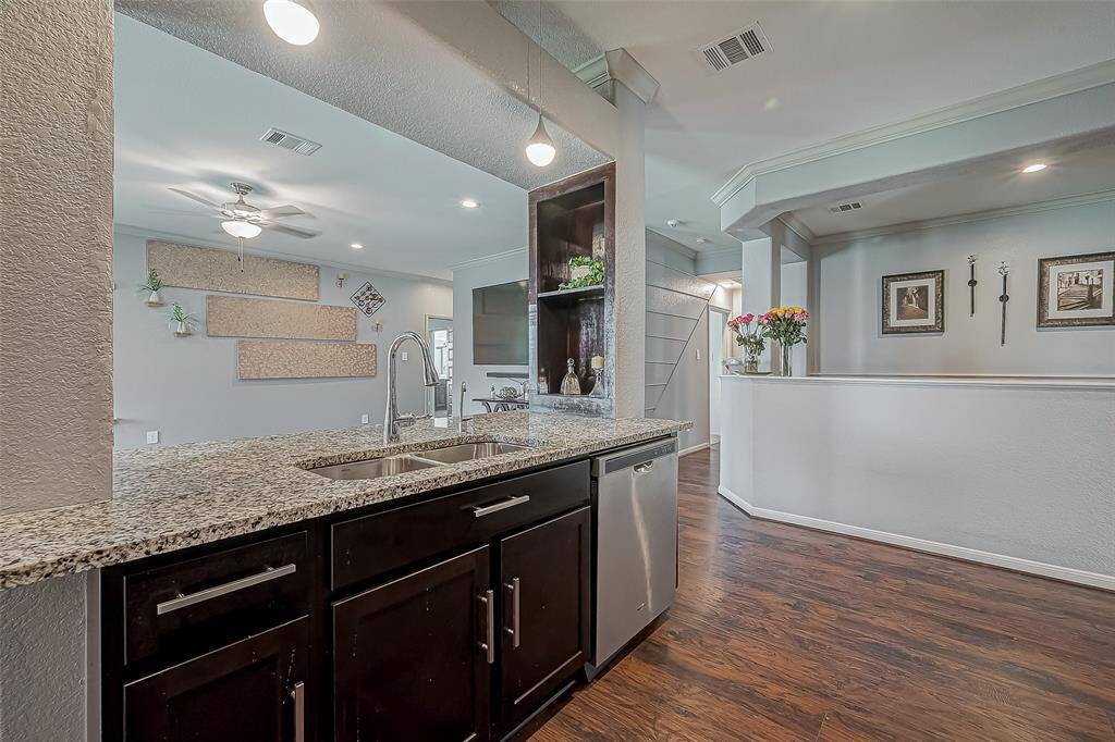This photo showcases a modern kitchen with granite countertops and a double sink. The space opens into a living area with laminate wood-like floors, recessed lighting, and a ceiling fan. The walls are adorned with decorative elements and framed art.