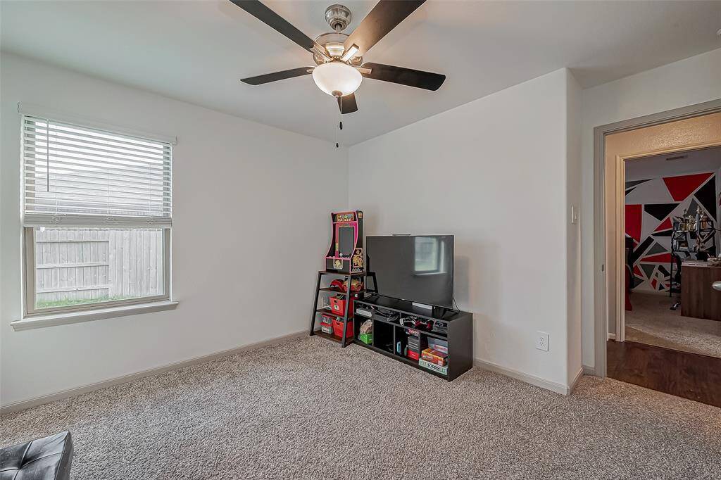 This cozy room features neutral carpeting, a ceiling fan with light, and ample natural light from a window overlooking a fenced yard.