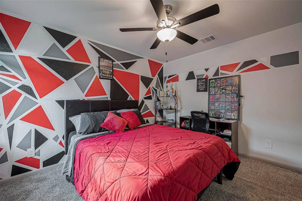Another view of the secondary bedroom featuring a bold geometric accent wall with red, black, and gray patterns. Notice the ceiling fan and soft carpeted flooring.