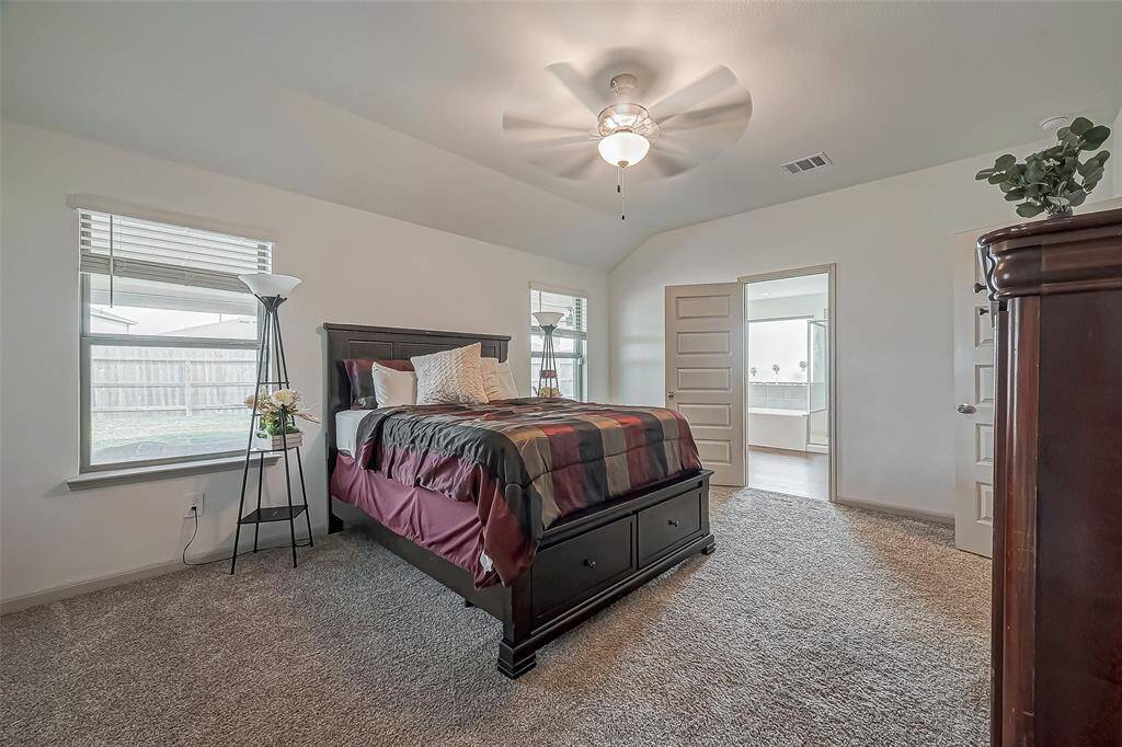 This beautiful primary bedroom features a cozy and spacious layout with carpet flooring, a ceiling fan, and ample natural light from two windows.