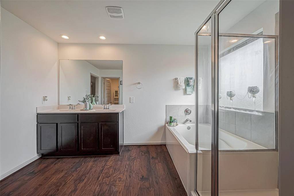 This bathroom features a modern design with a double vanity, large mirror, sleek glass-enclosed shower, and a separate bathtub. It has laminate wood-like flooring and ample natural light for a bright, inviting atmosphere.