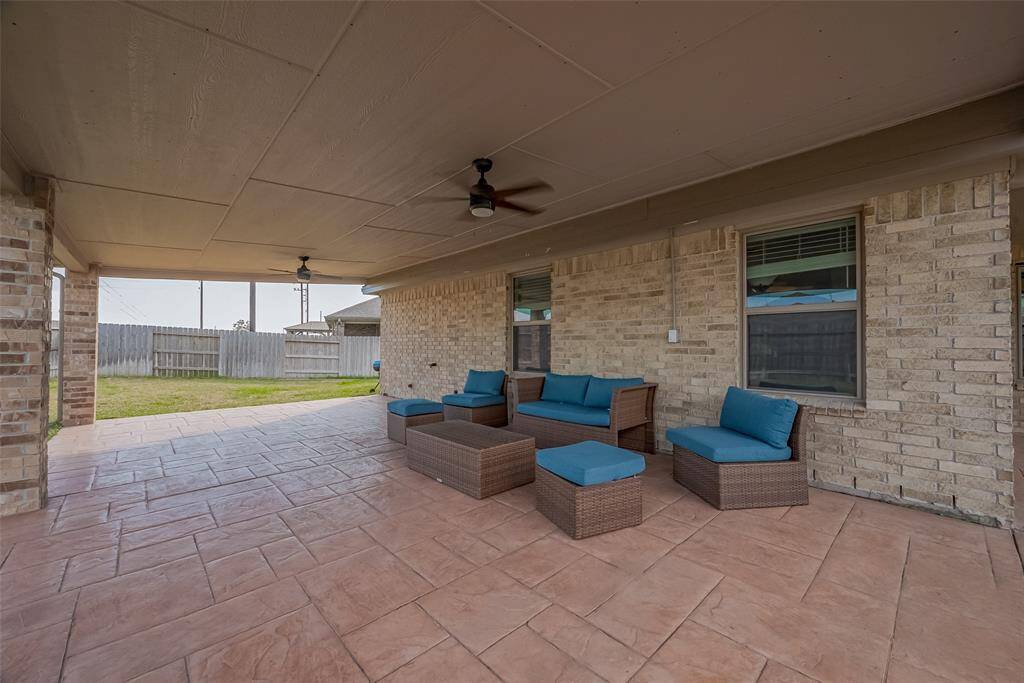 This photo showcases a spacious covered patio with stylish outdoor furniture and ceiling fans. The area is paved with decorative tiles and features brick walls, offering a comfortable and inviting space for relaxation and entertainment, with a view of a fenced backyard.
