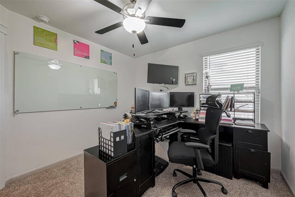 This photo shows a great home office/study space. The room features a ceiling fan, plush carpet and a window with blinds, providing a bright and efficient workspace.
