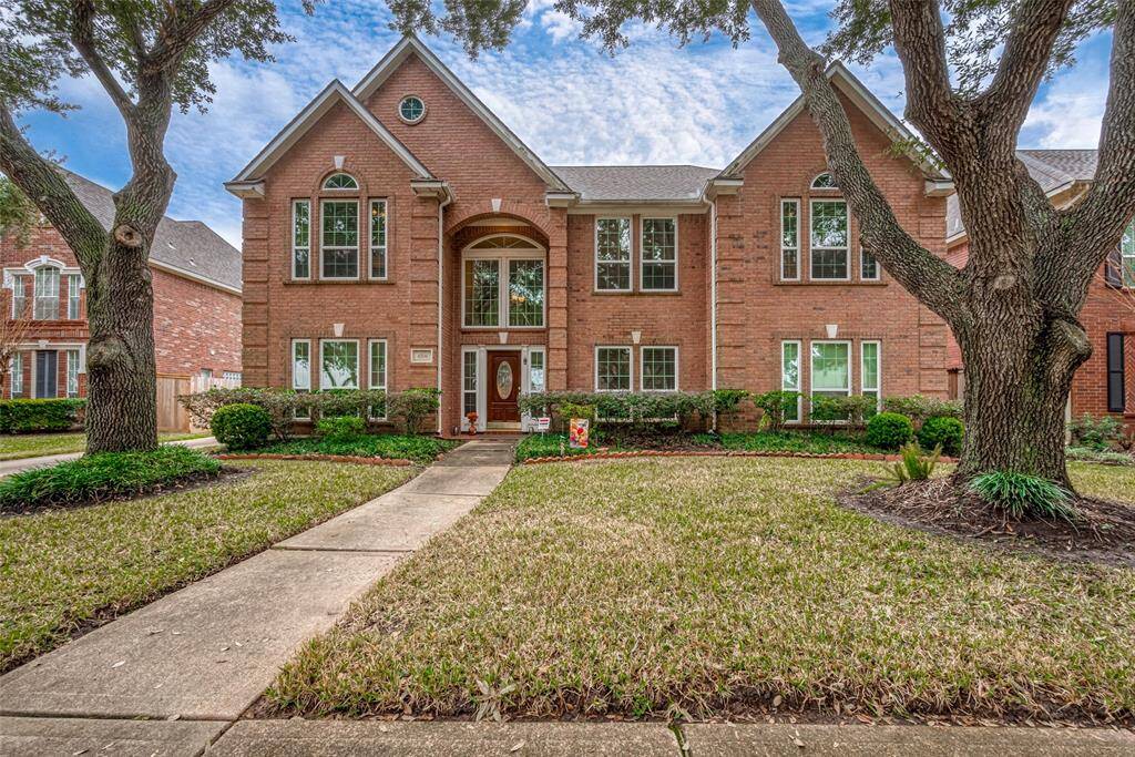 This photo showcases a beautiful two-story brick home with large windows and a welcoming entrance. The property features a well-maintained front yard with mature trees and a paved walkway leading to the front door.