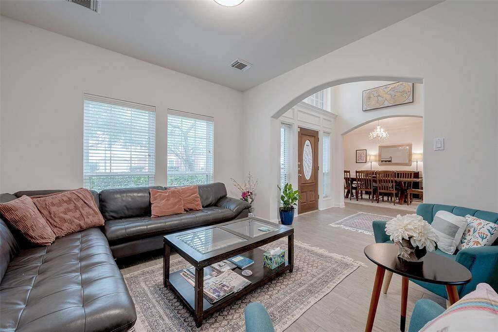 This cozy living room features a modern sectional sofa, a glass coffee table, and large windows that let in natural light. The open archway leads to a dining area with a chandelier, creating a welcoming and spacious feel. Neutral tones and decorative accents add warmth and style.