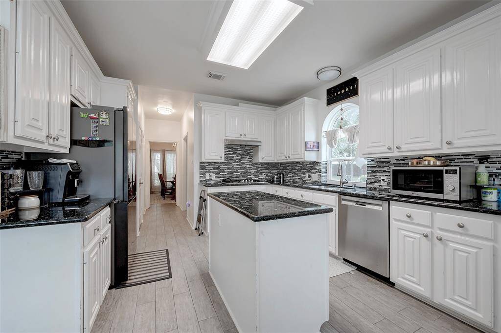 Bright, modern kitchen featuring white cabinetry, granite countertops, and a stylish tile backsplash. Includes a center island, stainless steel appliances, and ample natural light from a large window.