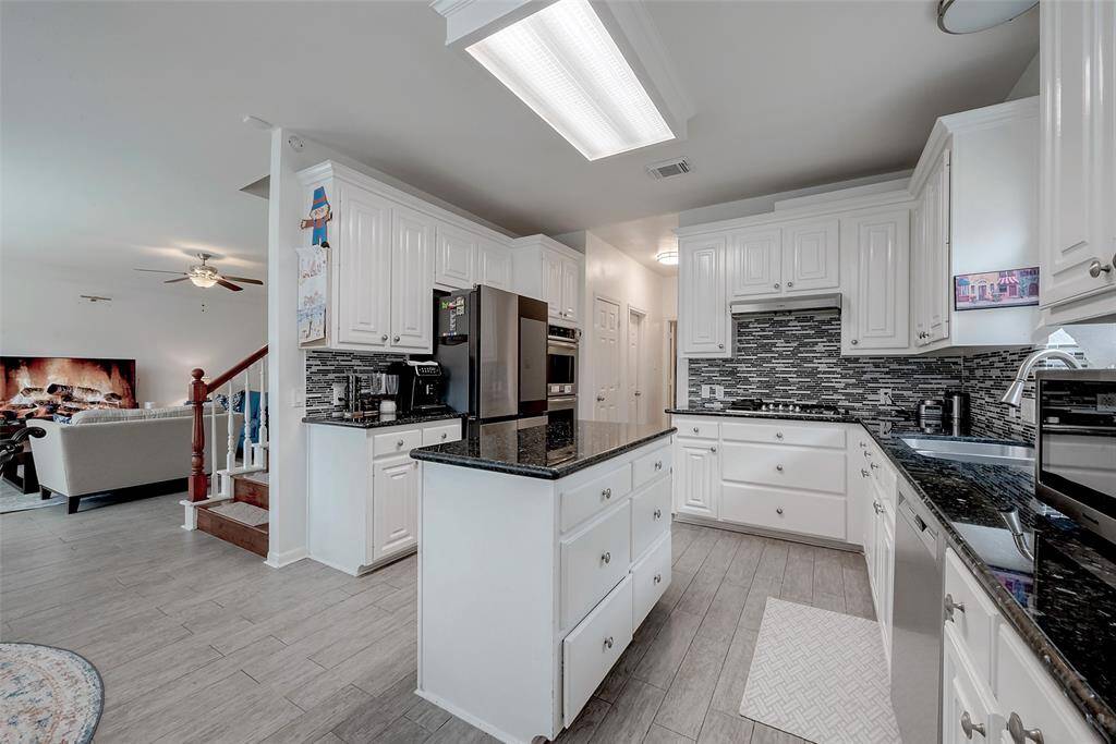 Bright, spacious kitchen with white cabinetry, granite countertops, and a modern tile backsplash. It features a central island, stainless steel appliances, and wood-look flooring. The kitchen opens to a cozy living area with a visible staircase, creating a welcoming, open-concept layout.