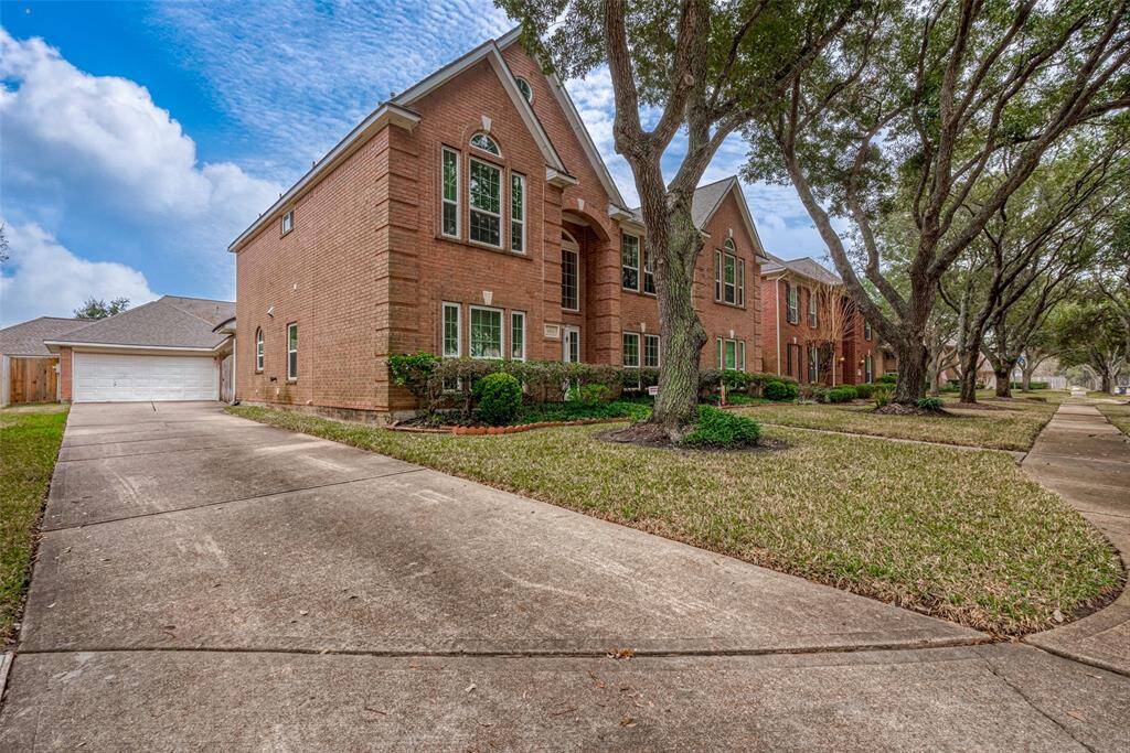 This photo features a spacious, two-story brick home with large windows and a well-maintained front yard. The house sits on a corner lot with mature trees lining the sidewalk. A long driveway leads to a detached two-car garage.