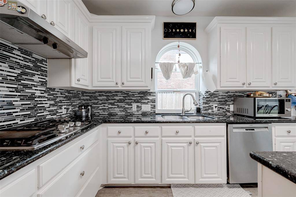 This kitchen features sleek white cabinetry with modern hardware, complemented by a stylish black and white tile backsplash. It includes a built-in gas stove, stainless steel dishwasher, and a dual-basin sink under a charming arched window, providing a bright and functional space.