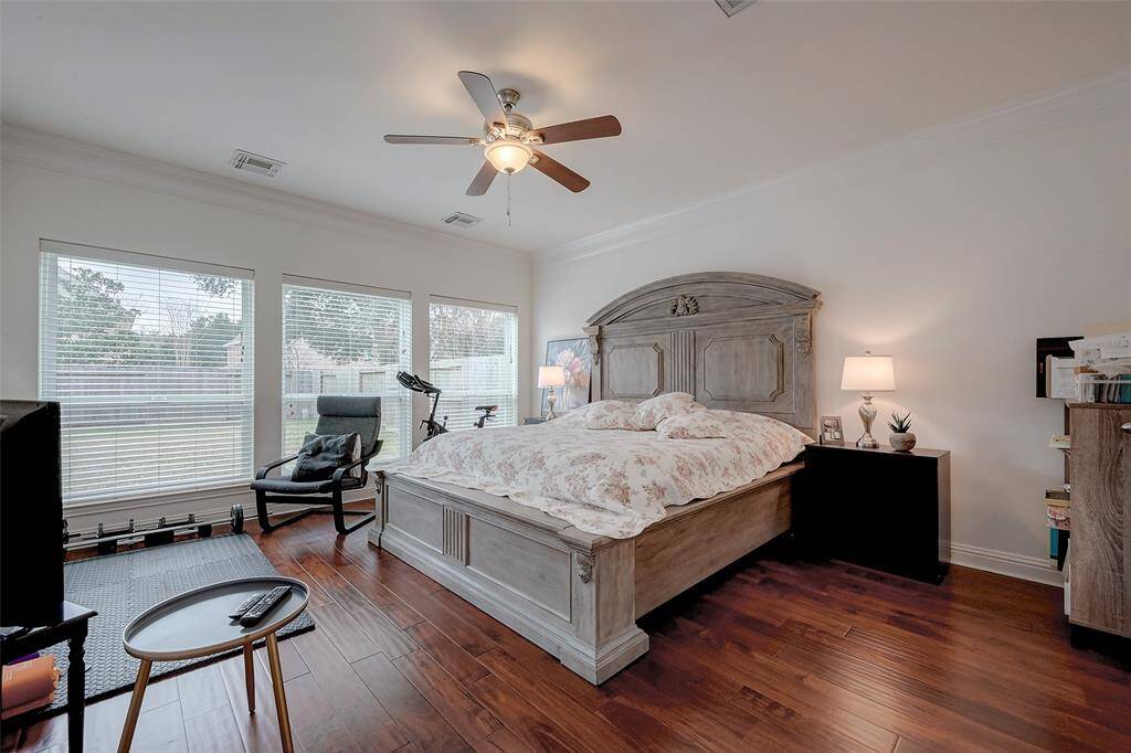 This spacious bedroom features rich hardwood floors and large windows that provide ample natural light. The room is equipped with a ceiling fan and includes a cozy seating area. It has a rustic wooden bed frame and nightstand, creating a warm and inviting atmosphere.