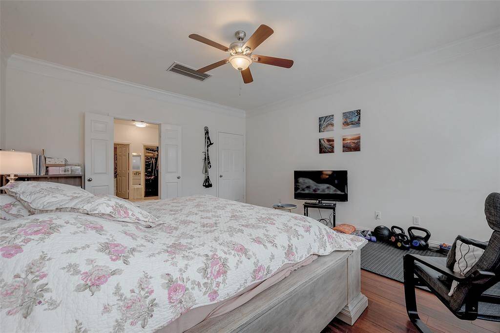 This spacious bedroom features a cozy atmosphere with a ceiling fan, wood flooring, and ample natural light. It includes a large bed with floral bedding, a TV setup, and a walk-in closet. The room is accented by wall art and offers a comfortable space for relaxation.