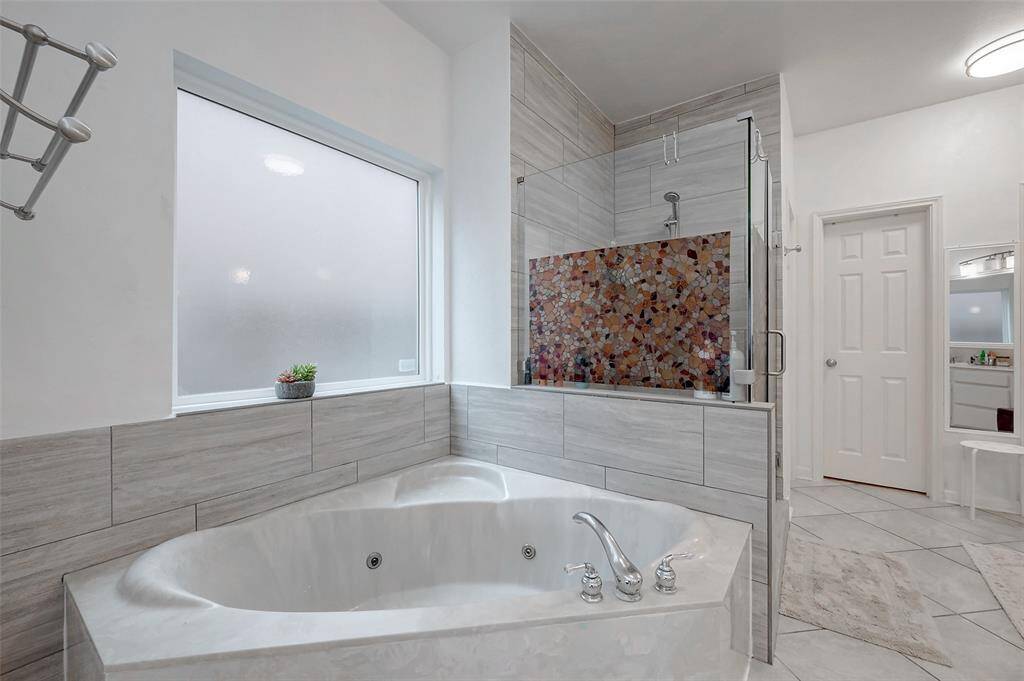 This bathroom features a luxurious corner soaking tub, a modern walk-in shower with a colorful mosaic accent, and ample natural light from a frosted window, creating a serene and elegant space.
