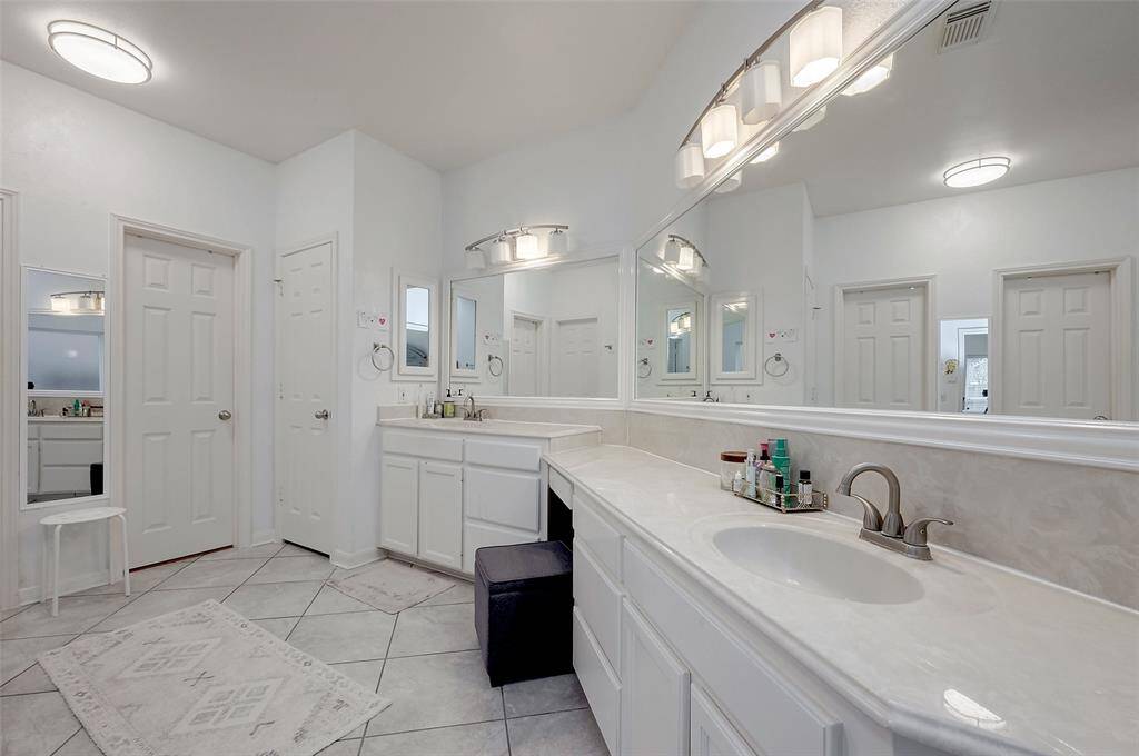 This spacious bathroom features dual sinks with a large mirror, ample cabinet storage, modern lighting, and a clean, neutral color palette. The tiled floor and additional seating add functionality and comfort.