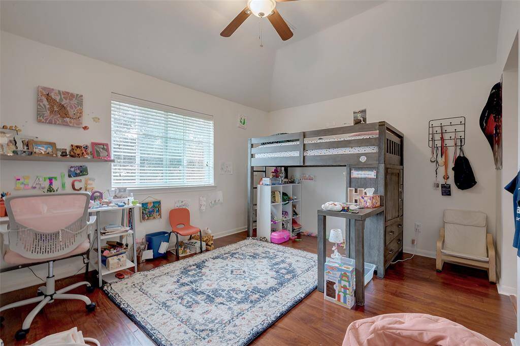 This cozy, well-lit children's bedroom features a loft bed with storage underneath, a spacious desk area, and a comfortable chair. The room has hardwood floors, a ceiling fan, and ample natural light from a large window, creating a welcoming space for study and play.