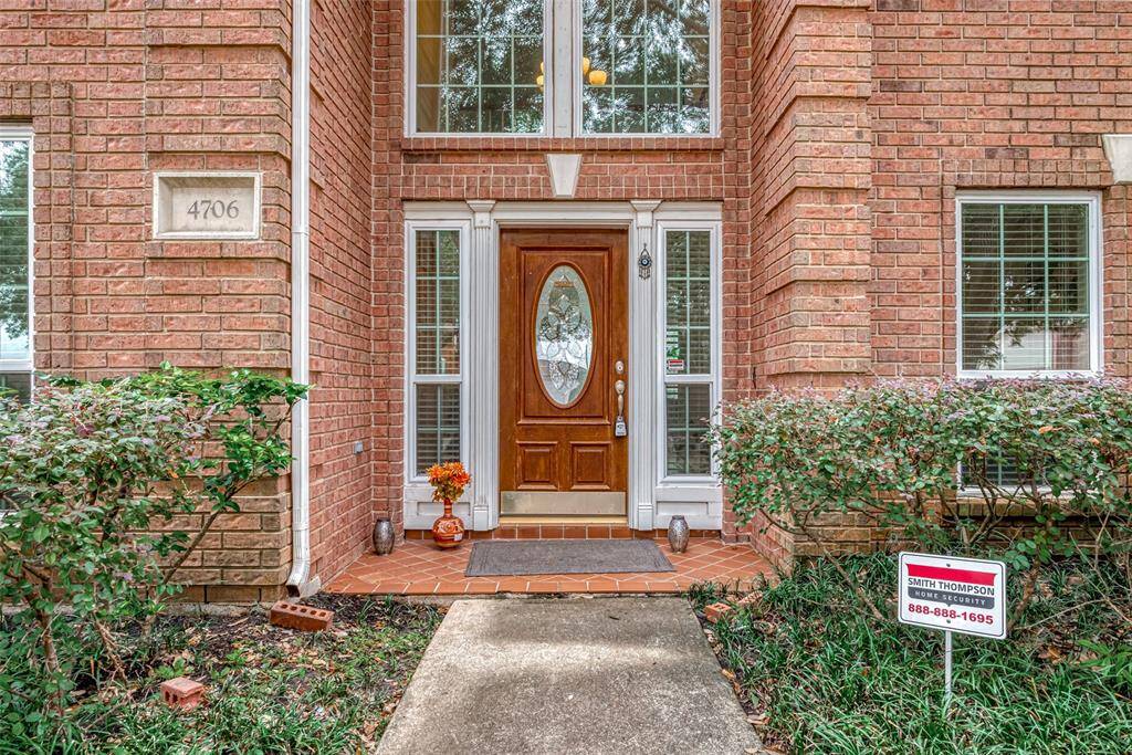 This inviting home entrance features a warm wood door with an oval glass insert, flanked by tall windows. Surrounded by lush greenery and a well-maintained brick exterior, it offers a welcoming and secure feel, highlighted by a visible home security sign.