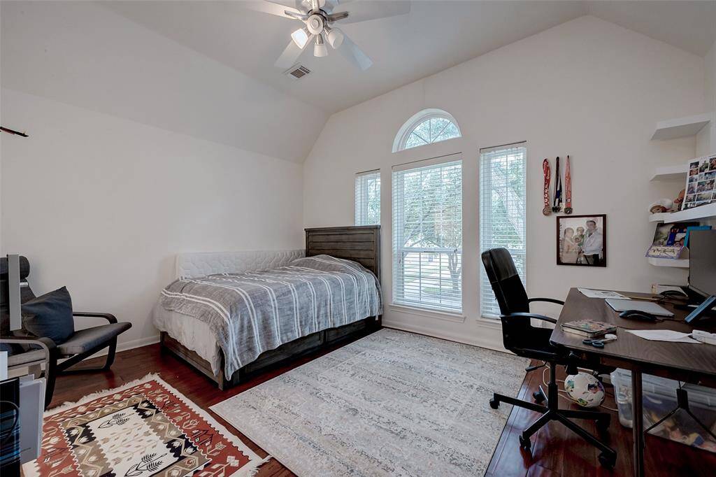 This photo showcases a bright, spacious bedroom with high ceilings and a ceiling fan. It features a cozy bed, a large window with an arched top that allows plenty of natural light, and a work area with a desk and shelves. The room has hardwood floors and a neutral color palette, creating a warm and inviting atmosphere.