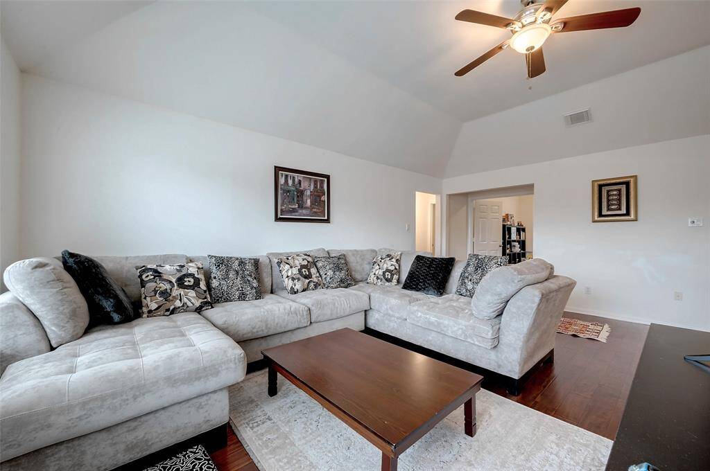 This photo showcases a spacious living room with a vaulted ceiling, featuring a large, comfortable sectional sofa and a wooden coffee table. The room is well-lit and decorated with wall art and a ceiling fan. The hardwood floors add warmth to the space, creating an inviting atmosphere.
