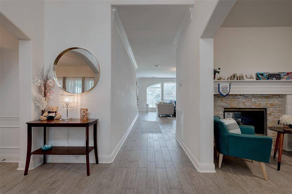 This photo showcases a bright, inviting hallway leading to a living area with large windows. The space is elegantly decorated, featuring a round mirror, a console table, and a cozy seating area by a stone fireplace. The flooring is a modern wood-look tile, enhancing the open and airy feel.