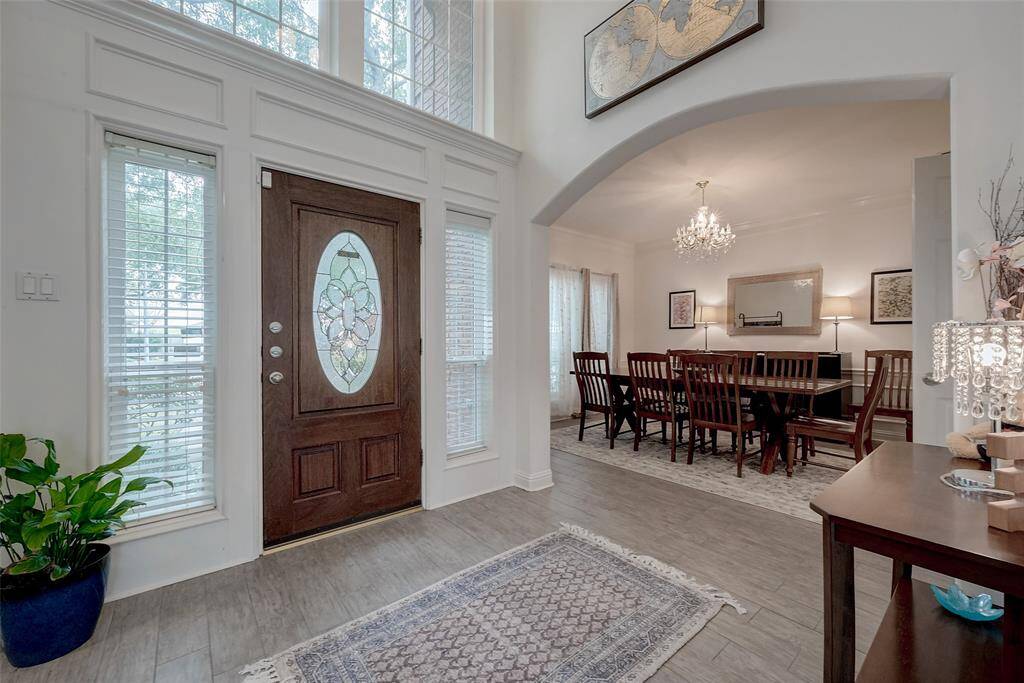 This inviting entryway features a stylish wooden front door with decorative glass, high ceilings, and ample natural light from tall windows. Adjacent is a formal dining room with elegant lighting and tasteful decor, creating a warm and welcoming atmosphere.