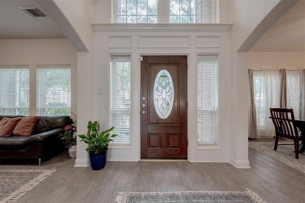 The photo showcases a welcoming entryway with a wooden door featuring an elegant oval glass design. Flanked by tall windows, the space is bright and airy. Adjacent rooms with a couch and dining set are visible, highlighting an open layout with light wood flooring and tasteful decor.