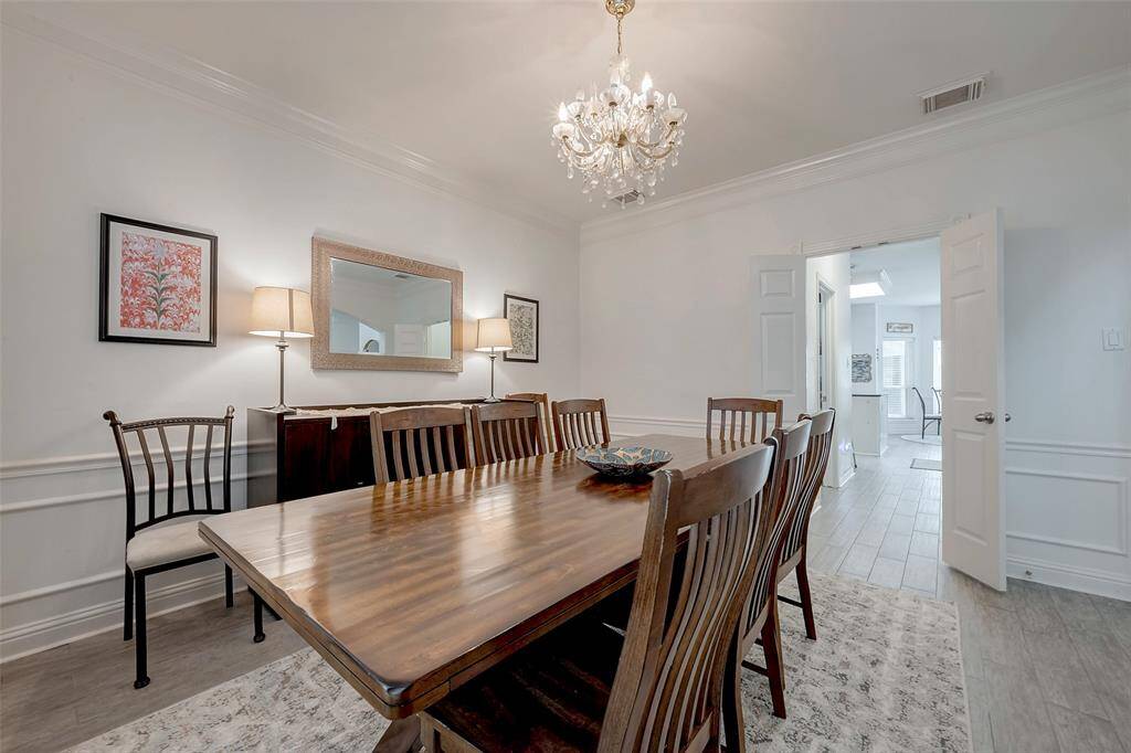 This dining room features a large wooden table with elegant chairs, a chandelier, and tasteful wall decor. It's bright and spacious, with white walls and wood flooring, creating a welcoming atmosphere.