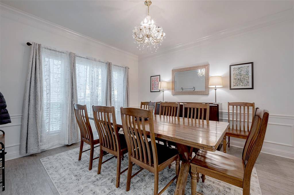This is a spacious dining room featuring a large wooden table with eight chairs, elegant chandelier lighting, and natural light streaming through sheer curtains. The room has a neutral color palette with decorative wall paneling and framed artwork, creating a warm and inviting atmosphere.