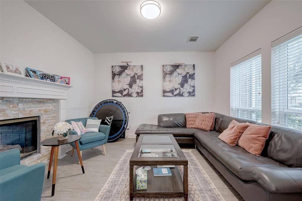 This cozy living room features a modern sectional sofa with accent pillows, a stone fireplace, and large windows for natural light. The decor includes floral artwork and a small trampoline, creating a family-friendly atmosphere.