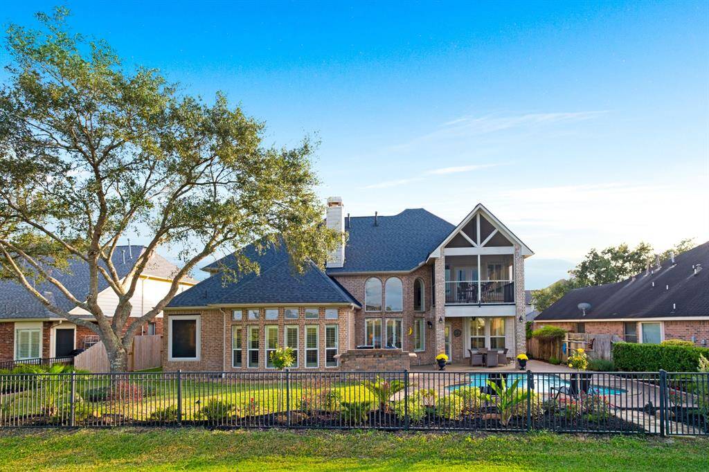 A spacious two-story brick home featuring a large balcony, a fenced backyard with mature trees, and a clear view of the sky above, suggesting a serene setting.