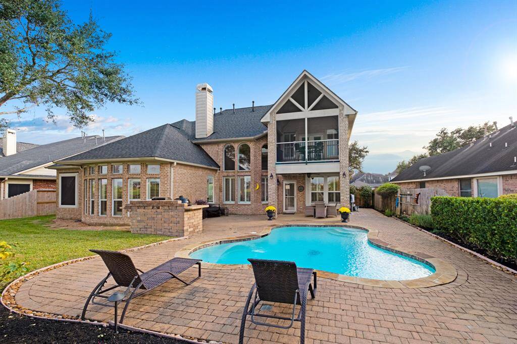 A spacious two-story brick home featuring a large in-ground pool in the backyard, with a cozy patio area and a well-maintained lawn. The house has ample windows, a prominent chimney, and a second-floor balcony overlooking the pool.