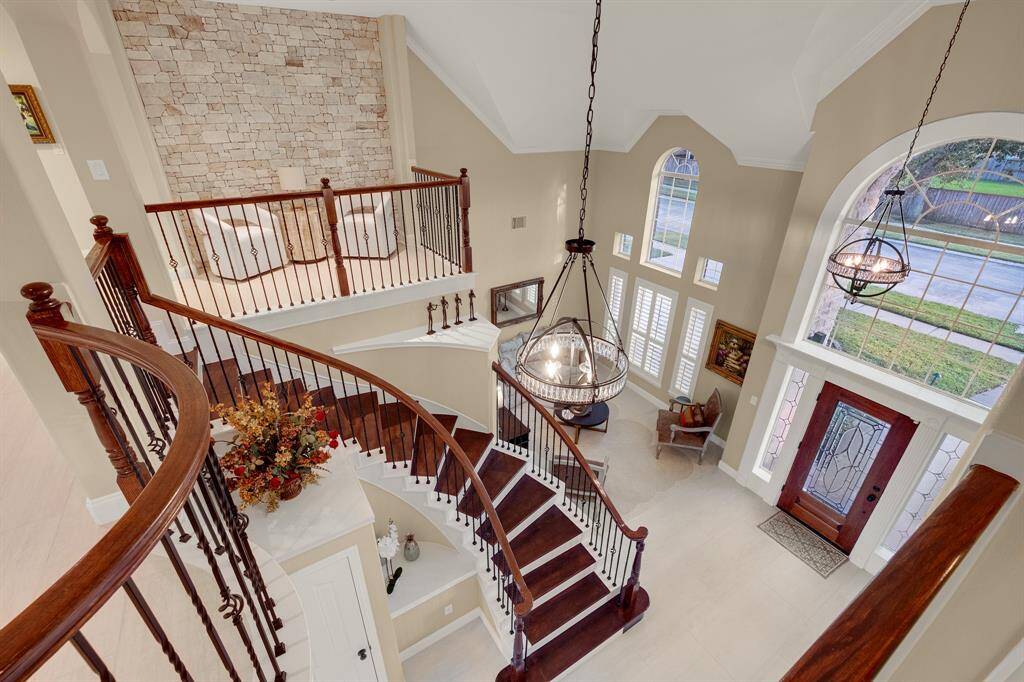 A spacious and elegant foyer featuring high ceilings, a grand staircase with wood and wrought iron railings, and ample natural light from large windows and an arched transom above the front door. The area is completed with a striking chandelier and a view of the second-floor landing.