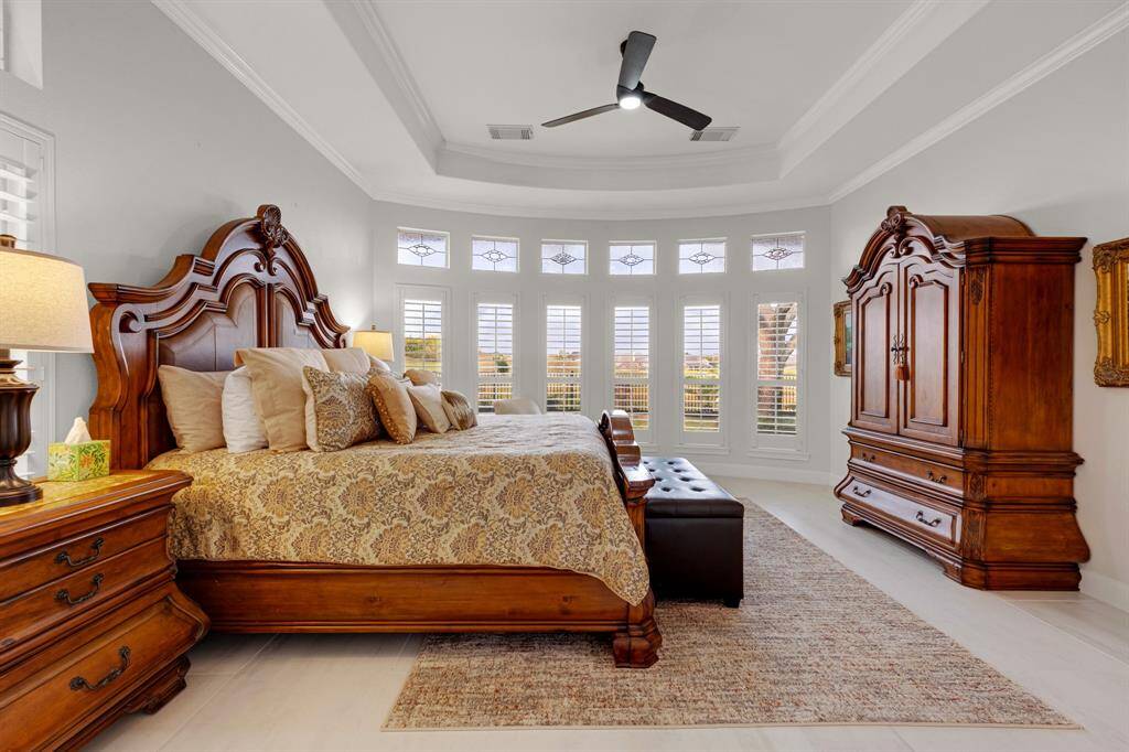 A spacious and elegantly appointed Primary bedroom. The room offers ample natural light through multiple windows adorned with plantation shutters and boasts a high tray ceiling with a ceiling fan.