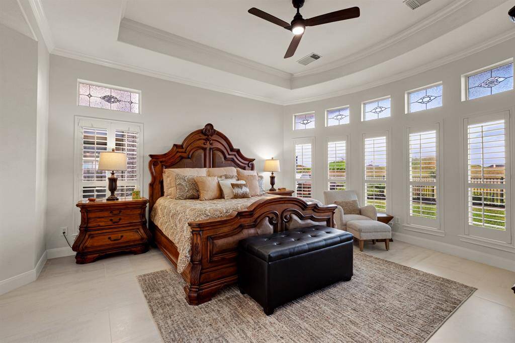 A spacious, well-lit bedroom and a sitting area with a view. The room includes a ceiling fan, elegant tray ceiling, and multiple windows with plantation shutters and decorative stained glass transoms, providing abundant natural light.