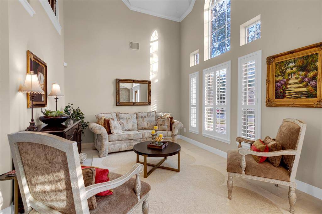 A spacious and well-lit living room featuring high ceilings, large windows with plantation shutters, and a neutral color scheme.