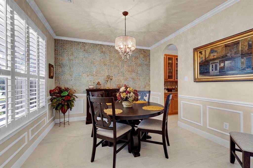 Elegant dining room featuring, stylish wainscoting, a unique tile accent wall, and large windows with plantation shutters, complemented by a classic chandelier and a view into the kitchen.
