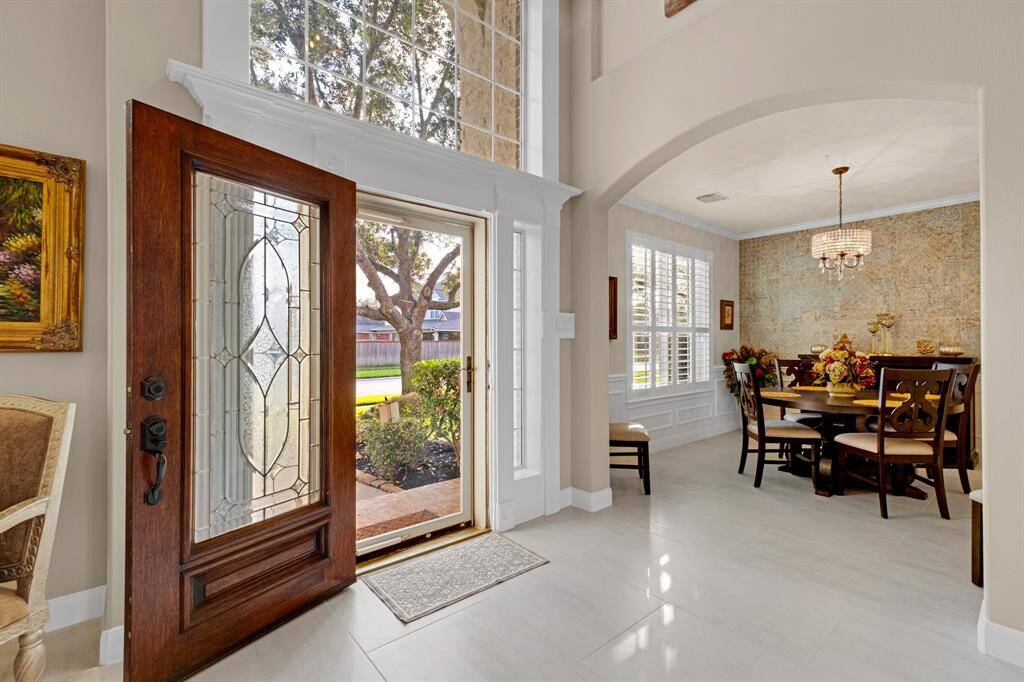 A bright and elegant dining area with a large window providing ample natural light, adjacent to an entryway featuring a stylish wood door with decorative glass. The space has high ceilings. * The entire lower level has new 2'x4' porcelain tile.